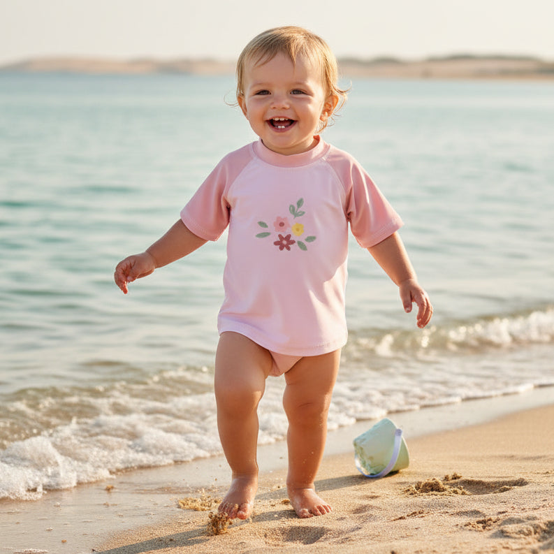 Baby trägt ein pinkes Bade-T-Shirt mit Blumenmotiv und läuft am Strand am Wasser entlang – sommerliche Baby-Bademode für Kinder