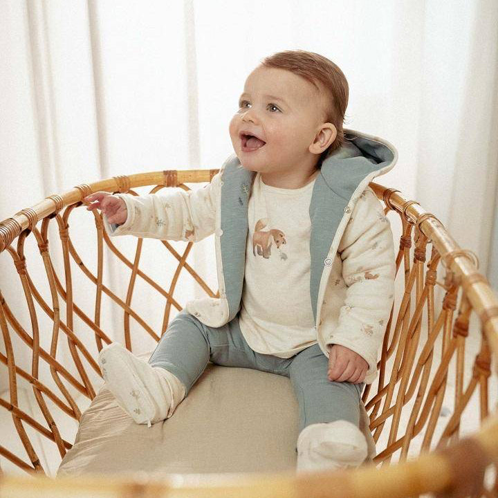 Baby sitzt in einem Korb und trägt eine weiche Wendejacke in Stone Blue – stilvolles Baby-Outfit, ideal als Geschenk zur Geburt