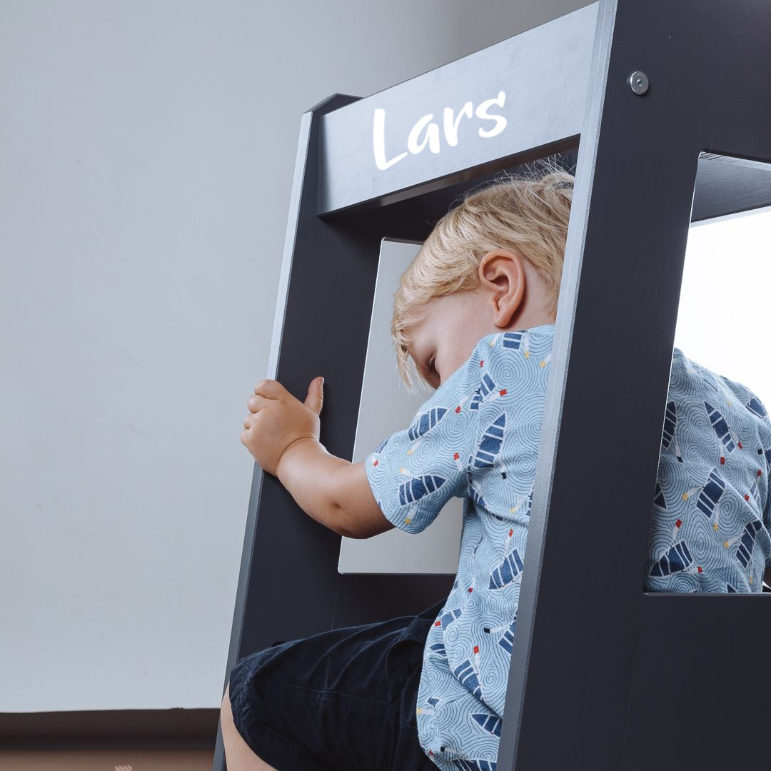 Child sitting on grey wooden learning tower with personalized name "Lars" visible on the top panel