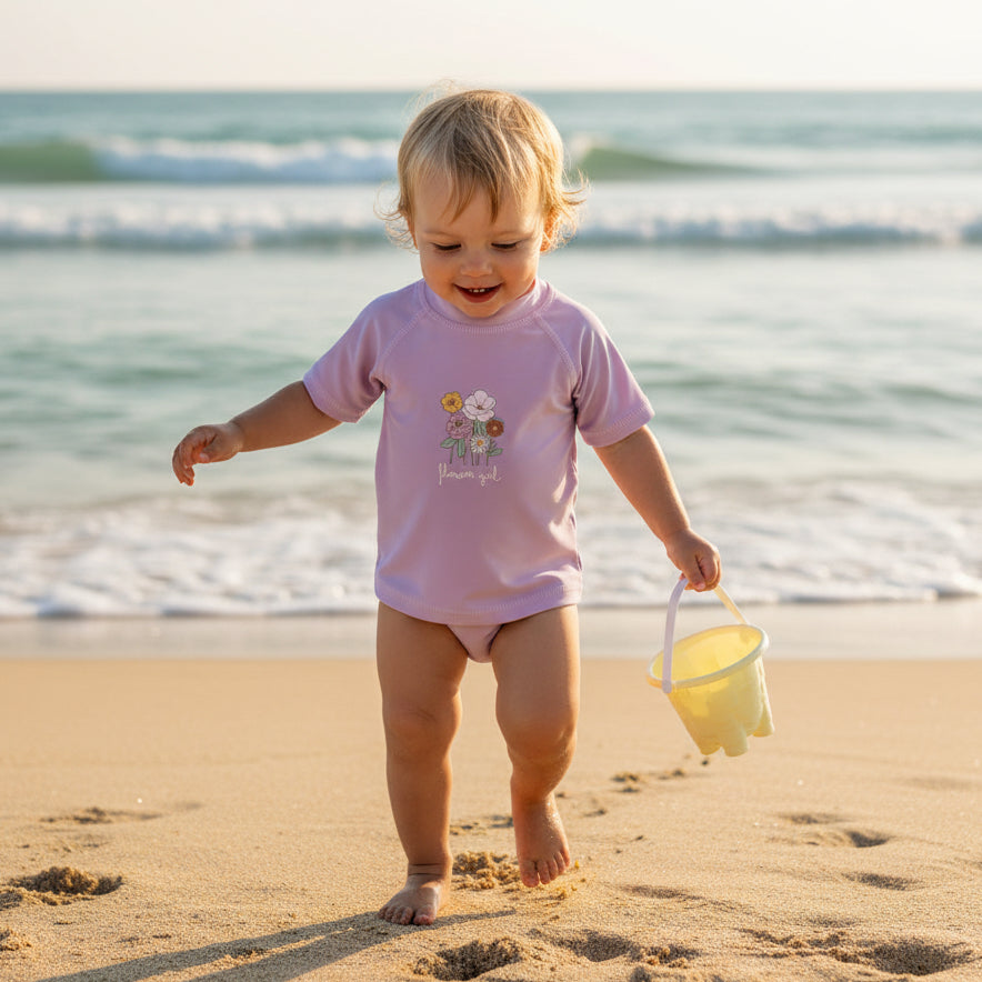 Baby läuft am Strand und trägt ein mauvefarbenes Bade-T-Shirt mit Blumenmotiv – sommerliche Baby-Bademode am Meer